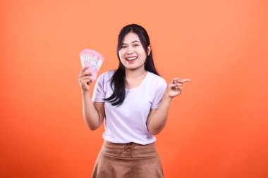 Elinde bir yığın Tayland Baht banknotu tutan ve turuncu arka planda izole bir şekilde sağ tarafı gösteren bir kadın..