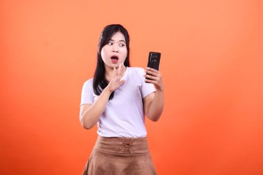 Şaşırmış genç bir kadın elinde bir akıllı telefon tutuyor ve diğer elini kaldırıyor. Turuncu bir arka planda izole edilmiş..