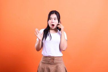Ağzı açık şaşırmış bir kadın turuncu arka planda kulağına bir cep telefonu tutuyor. Diğer elini kaldırdı. Beyaz bir tişörtü ve kahverengi bir eteği var..