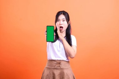 Şaşırmış genç bir kadın, turuncu arka planda izole edilmiş yeşil ekran ekranlı bir telefon tutuyor. Kadın beyaz bir gömlek ve kahverengi bir etek giyiyor ve eli açık ağzının yanında..