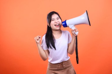 Bir kadın elinde megafonla turuncu bir arkaplanda izole edilmiş bir anons yapıyor. Uzun siyah saçları var ve beyaz bir tişört ve kahverengi bir etek giyiyor..