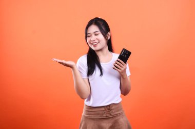 Gülümseyen bir kadın sağ elinde akıllı bir telefon tutar ve sol eliyle avucunu açar. Kadın turuncu bir arka planda izole edilmiş..