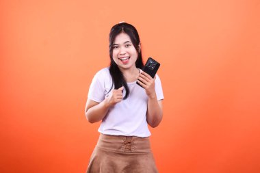 Gülümseyen bir kadın akıllı bir telefon tutuyor ve kameraya bakıyor. Beyaz bir gömlek ve kahverengi bir etek giyiyor ve turuncu bir arka planda izole edilmiş. Uzun siyah saçları var ve bir stüdyoda..