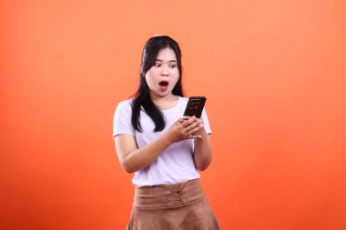 Siyah saçlı bir kadın ekranda şaşırmış bir ifadeyle siyah bir akıllı telefon tutuyor. O, beyaz bir tişört ve kahverengi bir etek giyiyor. Turuncu bir arka planda izole edilmiş..