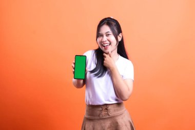Siyah saçlı bir kadın gülümsüyor ve elinde yeşil ekranlı bir akıllı telefon tutuyor. O, beyaz bir tişört ve kahverengi bir etek giyiyor. Arka plan turuncu. Kadın turuncu arka planda izole edilmiş..