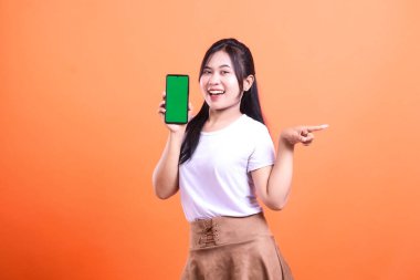 Beyaz tişört ve kahverengi etek giyen bir kadın yeşil ekranlı bir telefon tutuyor ve turuncu arka planda sırıtarak sağ tarafı işaret ediyor..