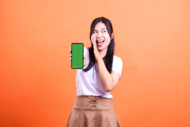 Genç bir kadın heyecanlı ve bir eli ağzı açık, bir eli ağzının kenarında turuncu bir arka planda izole edilmiş yeşil ekranlı akıllı bir telefon cihazı tutuyor.