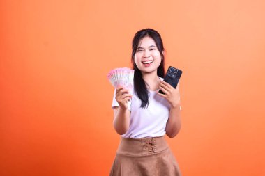 Siyah saçlı bir kadın bir elinde banknotlar, diğer elinde cep telefonu tutarken gülümsüyor. Beyaz bir gömlek ve kahverengi bir etek giyiyor ve turuncu bir arka planın karşısında duruyor..