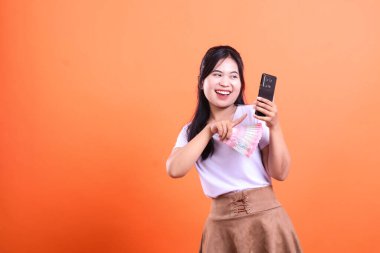 Neşeli bir kadın elinde bir yığın Endonezya rupia banknotu ve iki elinde de geniş bir gülümsemeyle siyah bir akıllı telefon aleti tutuyor. Arkaplan sade turuncu; turuncu arka planda izole..