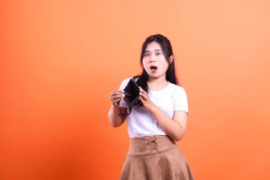 Yüzünde şok edici bir ifadeyle açık ve boş bir siyah cüzdan taşıyan bir kadının sade turuncu arka planda çekilmiş bir fotoğrafı..