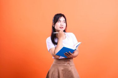 Beyaz tişörtlü ve kahverengi etekli genç bir kadın elinde mavi bir kitap ve bir kalem tutarken turuncu arka planda izole edilmiş tarafa bakıyor.