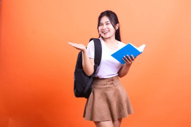 Siyah sırt çantalı genç bir kadın elinde mavi bir kitapla ayakta duruyor, diğer eli açık, beyaz bir bluz ve kahverengi bir etekle parlak turuncu bir arka plana gülümsüyor..