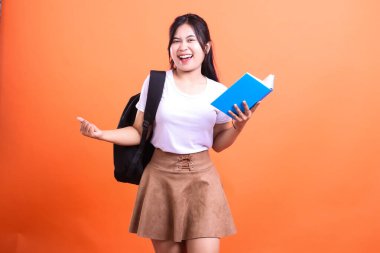 Sırt çantalı genç bir kadın turuncu arka planda mavi bir kitap tutuyor.