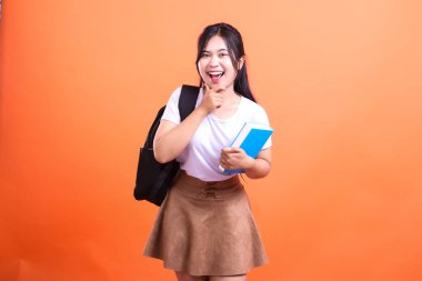 Siyah sırt çantası ve mavi kitabı olan genç bir kadın turuncu arka planda izole edilmiş bir stüdyo resmi için poz veriyor.