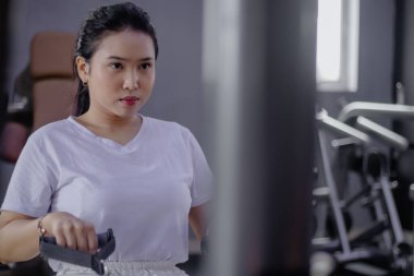 Azimli bir Asyalı kadın spor salonunda çekme makinesiyle egzersiz yapıyor. Sağlığa, sağlığa ve güce odaklan. Sahne, yoğun bir egzersiz anını yakalıyor..