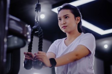 Odaklanmış genç bir kadın, trisepslerini salonda ip teçhizatıyla çalıştırıyor. Forma ve refaha modern ve kendini adamış bir yaklaşımı vurgular..