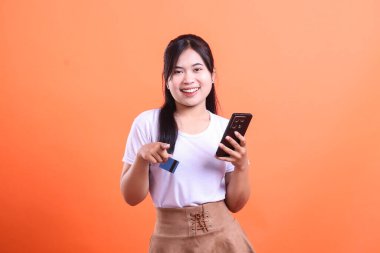 Genç bir kadın, elinde telefon ve kredi kartıyla online alışverişe hazır bir şekilde gülümsüyor. turuncu arkaplanda izole