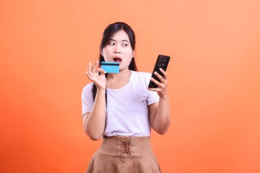 Genç bir kadın, elinde bir kredi kartı ve cep telefonu tutarken, internetten yapılan bir satın alma karşısında şaşkına dönüyor. turuncu arkaplanda izole