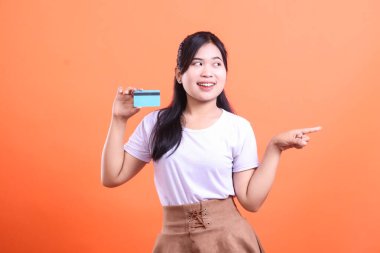 Elinde kredi kartıyla mutlu genç bir kadın, parmağını yana doğru gösteriyor, turuncu arka planda izole edilmiş heyecanlı görünüyor.