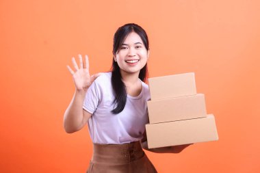 Elinde üç karton kutu tutan genç, neşeli bir kadın gülümsüyor ve turuncu arka planda izole bir şekilde kameraya el sallıyor..