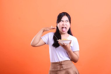 Genç bir kadın elinde bir kase erişte tutarken tiksindiğini ifade eder. Dilini uzatır ve elini turuncu arka planda izole bir şekilde yüzünü kapatır.