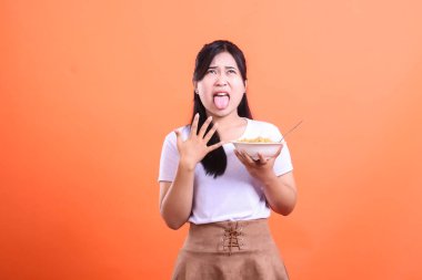 Genç bir Asyalı kadın yemek yedikten sonra tiksindiğini ifade ediyor, yüzü buruşmuş ve dili dışarı çıkmış, turuncu arka planda soyutlanmış.