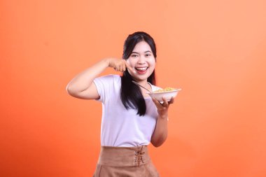 Genç bir kadın gülümsüyor ve diğer elinde bir kase erişteyle ağzını işaret ediyor. Yüzünde turuncu arka planda izole edilmiş neşeli bir ifade..