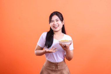 Gülümseyen genç bir kadın elinde bir kase erişteyle izleyiciye elini açarak sunuyor. turuncu arkaplanda izole