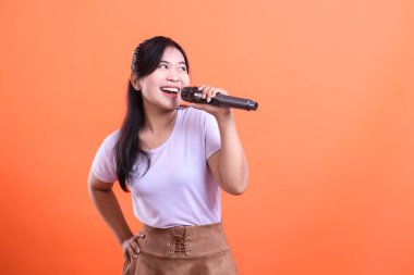 Neşeli bir Asyalı kadın mikrofonla şarkı söylüyor ve turuncu arka planda izole edilmiş mutlu bir ifadeyle yukarı bakıyor..