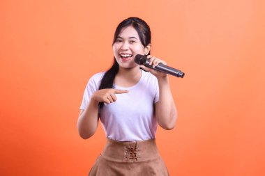 Mikrofonu işaret eden neşeli genç bir kadın, şarkı söylemeye hazır, ya da turuncu arka planda izole edilmiş, büyük bir gülümseme ile bir şov duyuran.