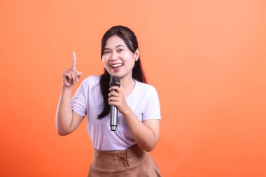 Genç bir kadın bir mikrofon tutuyor ve neşeli ve enerjik bir şekilde gülümserken parmağını havaya kaldırıyor. turuncu arkaplanda izole