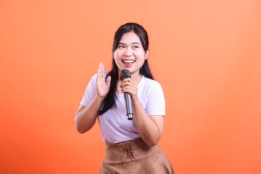 Genç bir kadın, mikrofonu tutarken ve elini sallarken mutlu bir şekilde gülümsüyor, sahnede şarkı söylemeye hazır, turuncu arka planda soyutlanmış..