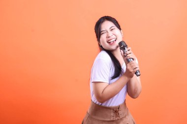 Genç ve anlamlı bir Asyalı kadın, gözleri kapalı ve turuncu arka planda izole bir şekilde mikrofona şarkı söylüyor..