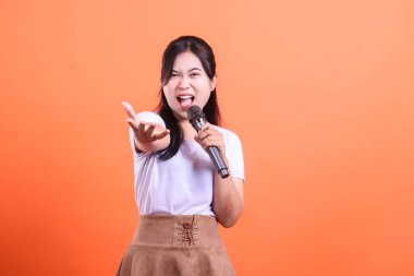 Elinde mikrofonla şarkı söyleyen hevesli genç bir kadın. Bize uzanan bir eli var, turuncu arka planda izole edilmiş.