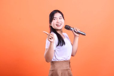 Gülümseyen genç bir kadın mikrofonla şarkı söylüyor, parmağıyla yan tarafı gösteriyor ve yukarı bakıyor, mutlu ve neşeli görünüyor, turuncu arka planda soyutlanmış.