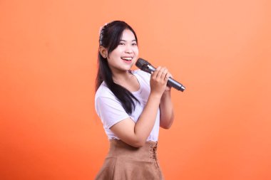 Hevesli genç bir kadın mikrofona şarkı söylüyor. Turuncu arka planda izole edilmiş müzik yeteneğini ve neşesini sergiliyor..