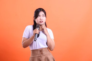 Genç bir kadın eli boynunda bir mikrofonla endişeli görünüyor. Sanki turuncu arka planda izole edilmiş bir performans ya da halka açık bir konuşma yapıyormuş gibi.