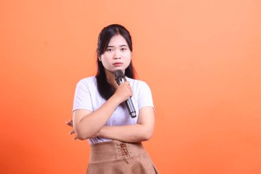 Ciddi bir genç kadın, turuncu arka planda izole edilmiş, tarafsız bir ifadeyle kameraya bakarken mikrofon tutuyor.