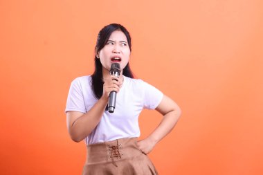 Genç bir kadın mikrofona anlamlı bir şekilde şarkı söylüyor, ciddi ya da esprili bir ifadeyle, turuncu arka planda izole edilmiş bir şekilde.