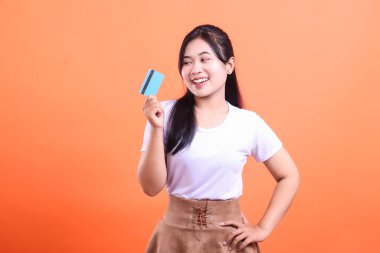 Genç bir kadın, bir eli belinde kredi kartı tutarken gülümsüyor, turuncu arka planda güven ve memnuniyet gösteriyor..