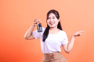 Neşeli Asyalı kadın bir araba anahtarı tutuyor ve gülümseyerek fotokopi alanını işaret ediyor. turuncu arkaplanda izole.