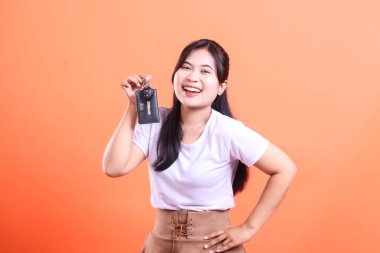 Heyecanlı genç bir kadın elinde kocaman bir gülümsemeyle araba anahtarı tutuyor. Turuncu arka planda izole edilmiş yeni bir araba hayali kuruyor.