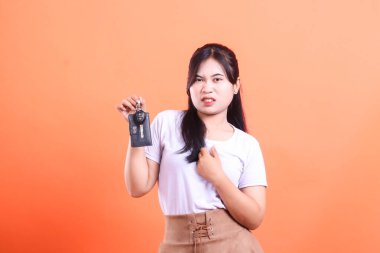 Asyalı bir kadın arabanın anahtarlarını tutarken tiksinti gösterir. Yüzü güçlü negatif duygular ve hoşnutsuzluk ifade eder. Turuncu arka planda izole edilmiş..