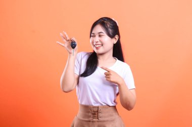 Mutlu genç bir kadın araba anahtarı tutuyor ve parmağıyla işaret ediyor, yeni araba sahipliğini veya ehliyetini kutluyor, turuncu arka planda izole edilmiş.