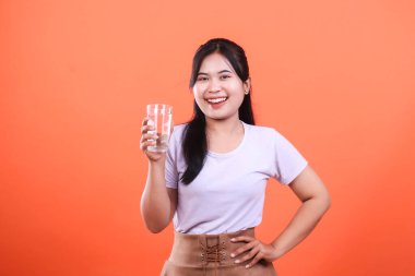 Gülümseyen Asyalı bir kadın elinde bir bardak su tutuyor, parlak turuncu arka planda sağlıklı su ve sağlığı teşvik ediyor. turuncu arkaplanda izole.