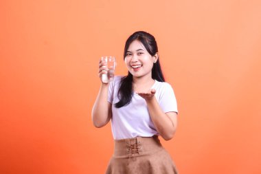 Elinde bir bardak su tutan ve eliyle el kol hareketi yapan gülümseyen genç bir Asyalı kadın susuzluğu ve sağlığı teşvik ediyor. turuncu arkaplanda izole