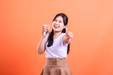 Genç Asyalı kadın gülümsüyor ve bir bardak su tutarken başparmağını kaldırıyor, turuncu arka planda izole edilmiş sağlıklı bir yaşam tarzını teşvik ediyor.