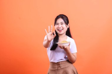 Neşeli genç bir kadın, elinde bir kase lezzetli erişte tutarken mükemmel bir jestle onayını veriyor. turuncu arkaplanda izole