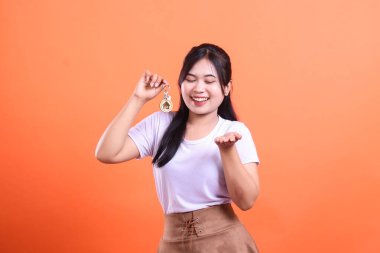 Gülümseyen genç bir kadın gururla sunar yeni bir evin anahtarlarını, açık bir avuç içi ile sanki anahtarları veriyor gibi, turuncu arka planda izole edilmiş.