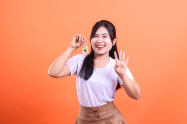 Mutlu genç Asyalı bir kadın yeni bir evin anahtarlarına sahip. Bu da ev sahipliği ve gayrimenkul başarısını simgeliyor. turuncu arkaplanda izole